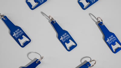 Diversos chaveiros de alumínio abridor garrafa com design em formato de garrafa na cor azul royal, personalizado com a frase "a sua gráfica, sempre",