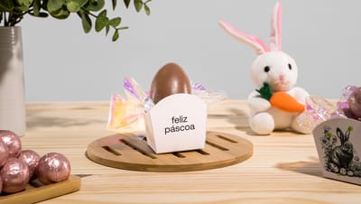 Embalagem cachepot personalizada com mensagem de "Feliz Páscoa",. Embalagem aberta mostrando um ovo de pascoa dentro, e com um coelho de pelúcia ao redor e ovos em cor rosê para compor a decoração