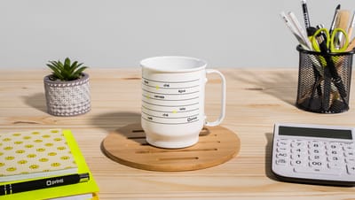 Caneca de alumínio branca apoiada em um suporte de madeira em uma mesa de escritório com itens de decoração ao redor, como calculadora, porta lápis, caderno e vasinho com planta