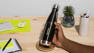 Garrafa térmica personalizada preta com acabamento metálico e tampa de aço inox, exibindo o logo da Printi, sendo segurada por uma mão sobre uma mesa de madeira. Ao redor, aparecem itens de escritório como bloco de notas com espiral, caneta, teclado, mous