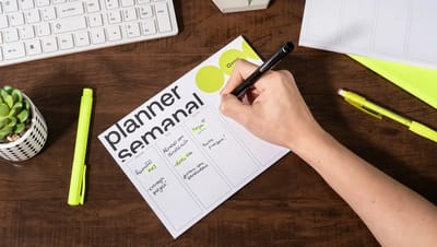 Planner semanal personalizado sobre mesa de madeira sendo preenchido à mão com canetas coloridas.