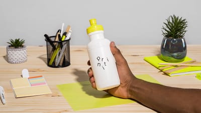 Squeeze plástico personalizado branco com tampa esportiva amarela, exibindo o nome Printi em tipografia minimalista. A garrafa está sendo segurada por uma mão sobre uma mesa de madeira com itens de escritório ao redor, como porta-canetas, bloco de notas, 