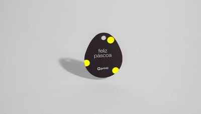 Tag para Ovo de Páscoa individual no formato de ovo, com fundo preto. O design inclui bolinhas amarelas posicionadas nos cantos e o texto “feliz páscoa” em branco centralizado, acompanhado da marca Printi na parte inferior.