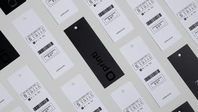 Conjunto de tags para roupas personalizadas dispostas sobre fundo claro, com etiquetas brancas exibindo informações de tamanho, código de barras e preço, além de tags pretas com o logo “printi”, ilustrando etiquetas de vestuário usadas para identificação 