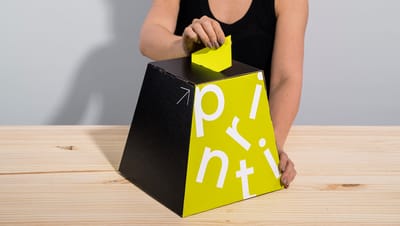 Pessoa usando urna de papelão da Printi com recorte superior para inserção de votos ou cupons