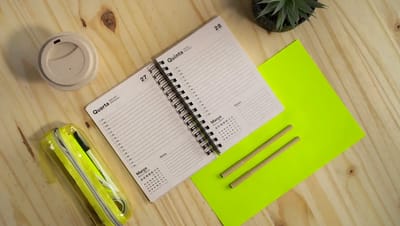 Agenda diária ecológica personalizada com encadernação espiral aberta sobre mesa de madeira, mostrando páginas de planejamento com linhas para anotações, ao lado de estojo transparente, canetas e copo reutilizável.