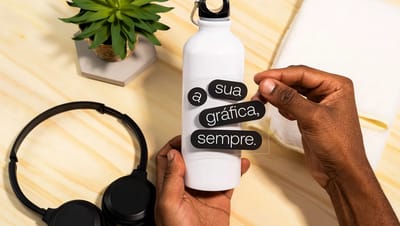 Pessoa aplicando adesivo quadrado transparente personalizado em garrafa branca, demonstrando a versatilidade de uso em brindes e acessórios de escritório.