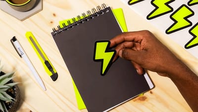 Mão aplicando adesivo personalizado de raio em caderno preto, demonstrando a aderência e o uso de papelaria personalizada em escritório.