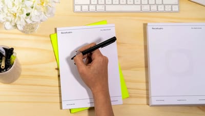 Visão superior de mão escrevendo em bloco de notas personalizado sobre mesa de madeira, com teclado de computador e flores ao fundo.