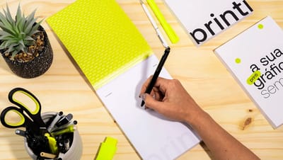 Pessoa escrevendo em um Bloco de Notas com Capa Blocado personalizado sobre uma mesa de madeira com materiais de escritório e um vaso de planta.