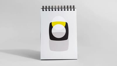 Bloco de notas com capa wire-o posicionado verticalmente, apresentando design minimalista com formas geométricas em preto e amarelo sobre fundo cinza claro.