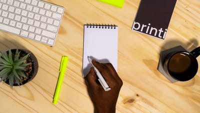 Pessoa escrevendo em um bloco para rascunho espiral branco sobre mesa de madeira com teclado, café e planta ao lado.
