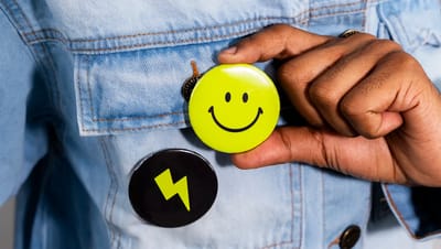 Botton personalizado redondo em amarelo neon com face sorridente sendo colocado em jaqueta jeans.
