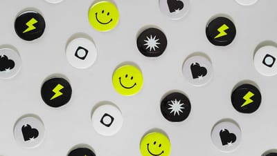 Composição de diversos bottons personalizados redondos com logotipos e ícones variados em preto, branco e amarelo.