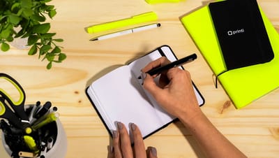 Mãos escrevendo em uma caderneta pequena aberta sobre mesa de madeira, cercada por itens de papelaria personalizados em preto e amarelo neon.