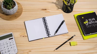 Caderno pequeno personalizado da Printi com capa impressa em design geométrico preto e branco, aberto sobre uma mesa ao lado de uma caneta plástica verde, destacando as páginas pautadas e o acabamento wire-o preto.