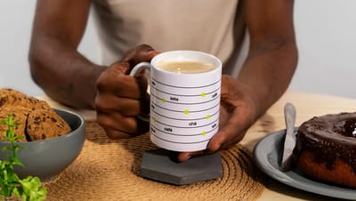 Pessoa segurando caneca personalizada da Printi com café em mesa de café da manhã, destacando o design criativo com níveis de bebidas.