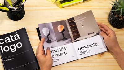 Mãos folheando catálogo personalizado da Printi sobre mesa de madeira, exibindo layout interno com fotos de luminárias e acabamento premium.
