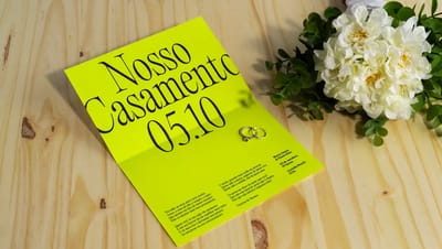 Convite de casamento moderno em papel verde-limão neon aberto sobre mesa de madeira, exibindo poema de Vinicius de Moraes e detalhes do evento ao lado de duas alianças e um buquê de flores brancas.