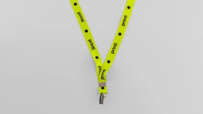 Cordão para crachá na cor verde, dobrado em forma de 'V', sobre uma superfície cinza neutra. A fita tem a marca 'Printi' impressa em preto com pontos pretos
