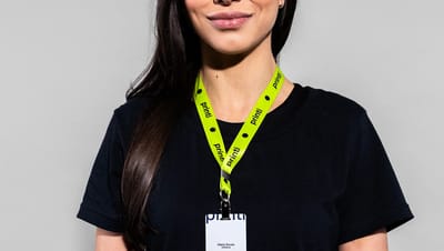 Mulher sorrindo, vestindo uma camiseta preta e um cordão para crachá verde neon. O cordão exibe o logo preto da Printi repetido e está preso a um crachá de identificação. O fundo é cinza claro.