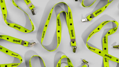 Várias opções de cordão para crachá personalizado na cor verde limão com a logo da Printi, espalhados sobre uma superfície cinza clara, destacando o acabamento com fecho tipo jacaré.