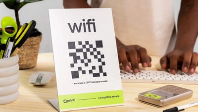 Display em L personalizado de mesa com QR Code para Wi-Fi da Printi, posicionado em cenário de escritório moderno com pessoa ao fundo digitando no teclado.