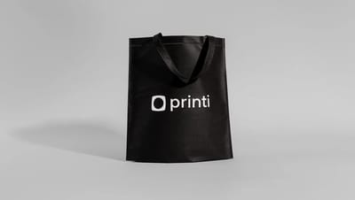 Ecobag personalizada de TNT preta com o logo da Printi centralizado, em pé contra um fundo cinza.