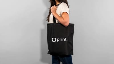 Mulher usando uma ecobag personalizada preta da Printi no ombro contra uma parede cinza.