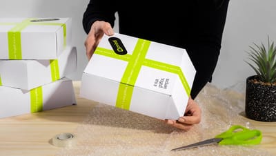 Pessoa manuseando caixa basculante personalizada da Printi em mesa de expedição com plástico bolha, evidenciando o uso prático para envios.