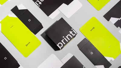 Foto com vista superior de diversas embalagens de papel desmontadas e uma montada da marca Printi. O design utiliza um contraste forte entre preto, verde-limão vibrante e branco. A caixa central, já montada, é preta com o logotipo "printi" em letras branc