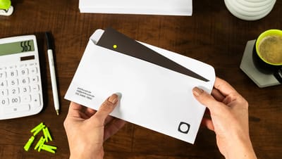 Mãos segurando envelope personalizado branco da Printi sendo aberto sobre uma mesa de madeira. A cena de escritório inclui calculadora, caneta, clips verdes e uma xícara de café, destacando o uso profissional do material.