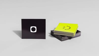 Um ímã de geladeira quadrado preto com logotipo centralizado ao lado de uma pilha de ímãs decorativos com estampa de carinha sorridente, exibidos em fundo cinza claro.