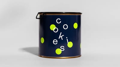 Lata de metal cilíndrica com design moderno na cor azul-marinho, estampa de bolinhas verdes e a palavra cookies, com tampa preta fechada em fundo cinza claro.