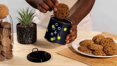Lata de metal para cookies azul-marinho decorada com bolinhas verdes, sendo segurada por uma pessoa que retira um cookie de chocolate de dentro dela, em uma mesa com prato de biscoitos e planta decorativa.