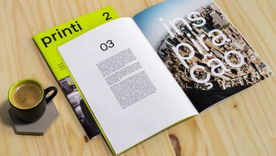 Livreto personalizado da Printi aberto em mesa de madeira ao lado de xícara de café, exibindo layout interno com textos e fotografia urbana de alta resolução.