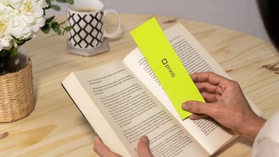 Pessoa utilizando um marca-página personalizado verde neon em um livro aberto sobre mesa de madeira, demonstrando o uso prático do produto.