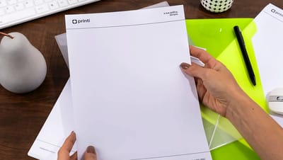 Mãos segurando folha de papel timbrado personalizado da Printi sobre mesa de escritório com caneta e pasta verde.