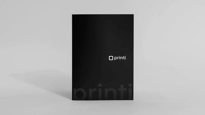 Pasta personalizada da Printi em papel preto fosco com acabamento premium. O design apresenta o logotipo centralizado em branco e uma marca d'água discreta da marca na parte inferior, sobre um fundo neutro.