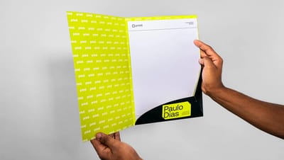 Mãos segurando pasta personalizada aberta com bolsa interna preta e porta-cartão. O interior é decorado com padrão de logotipos da Printi em fundo verde-limão e contém folhas brancas de papel timbrado.