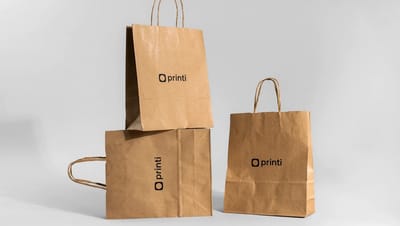 Três sacolas de papel kraft personalizadas empilhadas em fundo cinza claro, destacando o design, a resistência e o tamanho da embalagem corporativa.