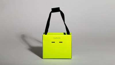 Sacola de papel personalizada modelo teepee em verde neon com estampa de ícone sorridente (smile) e alças pretas em fundo cinza neutro.