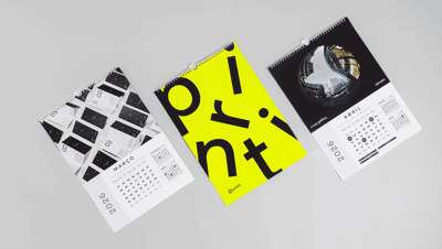 Calendário de parede premium personalizado com espiral metálica e suporte para pendurar, exibindo três páginas com layouts modernos, fotografia arquitetônica e design tipográfico em destaque, com visual clean sobre fundo neutro.
