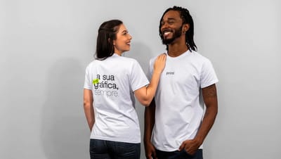Duas pessoas usando camisetas personalizadas brancas da Printi, em fundo neutro. Um homem veste camiseta com logo “printi” no peito, enquanto uma mulher mostra o verso da camiseta com a frase “a sua gráfica, sempre.” destacando o potencial de camisetas pe