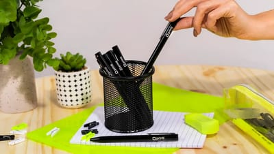 Mão retirando caneta metálica personalizada com logo Printi de um porta-canetas preto sobre mesa de madeira, cercada por itens de papelaria como marca-texto, clips, caderno pautado e estojo transparente, além de plantas decorativas, representando uma cane