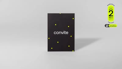 Foto de estúdio de um convite personalizado com fundo preto, a palavra "convite" centralizada em branco e pequenas bolinhas amarelas espalhadas. Ao fundo, uma parede cinza clara com um selo de entrega rápida.