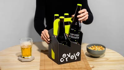 Embalagem personalizada para cerveja artesanal em papel cartão preto com alça verde neon, contendo garrafas de cerveja sendo retiradas por uma pessoa sobre uma mesa de madeira, ao lado de um copo de cerveja e petiscos