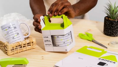 Embalagem personalizada para caneca sendo montada em mesa de trabalho, com design em branco e verde neon da Printi, ideal para presentear, proteger e valorizar canecas personalizadas em brindes ou vendas.