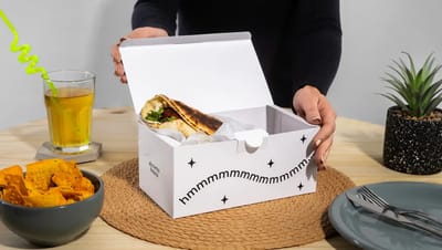 Pessoa abrindo embalagem personalizada para combos de comida, contendo sanduíche envolto em papel sobre mesa de madeira. Ao redor aparecem bebida com canudo, nachos, prato com talheres e planta decorativa