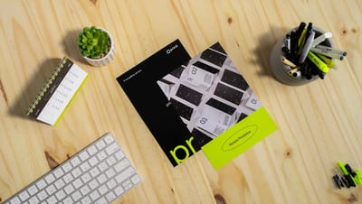 Flyer personalizado da Printi sobre mesa de madeira com teclado, calendário e acessórios de escritório, destacando novos produtos e identidade visual em preto e verde neon.